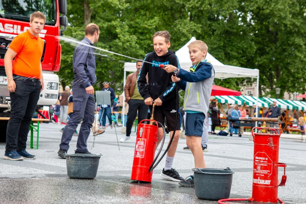 Dečka škropita vodo s pomočjo gasilske brizgalke v okviru delavnice na festivalu »Igraj se z mano«. Foto: Maša Pelc